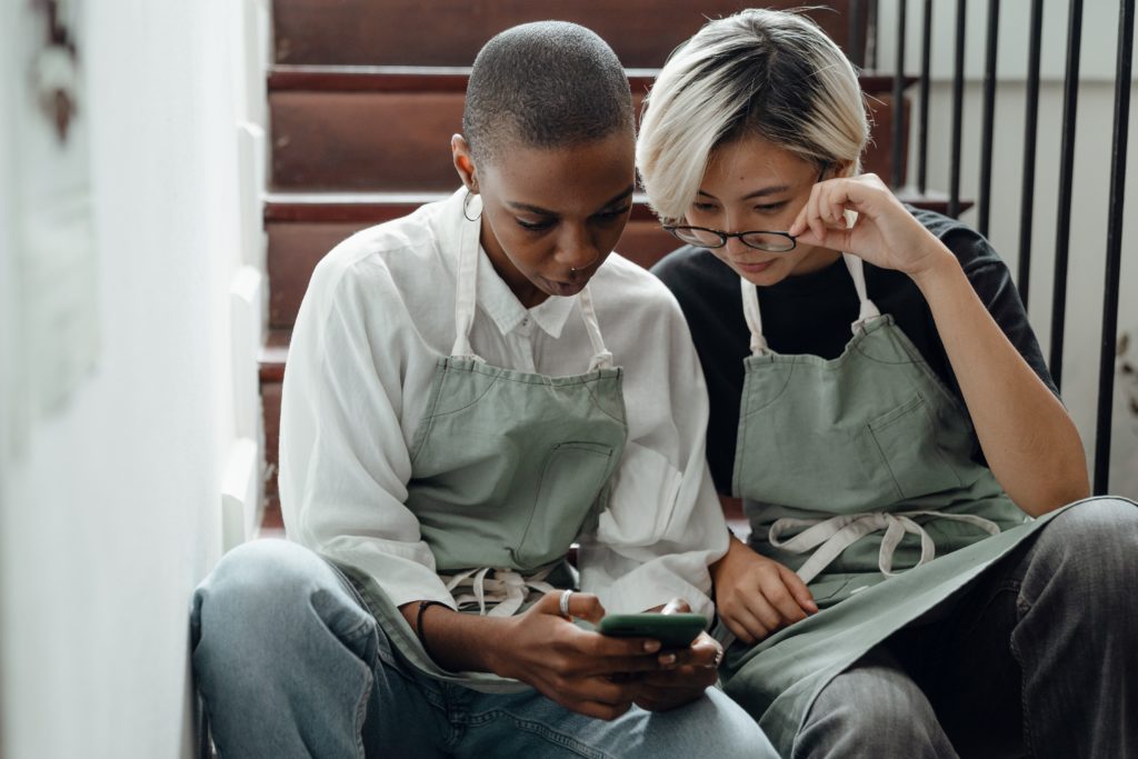 2 people sit on a brown staircase. One lowers glasses to view phone in the others hand. Both wear aprons
