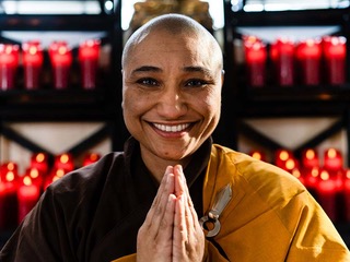 Sister Clear Grace smiling with palms together under chin. The background is two shelves of lit, tall, red candles.