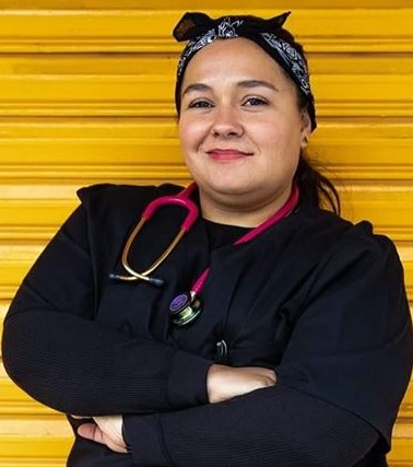 A waist up photo of a light skinned Mexican woman. She is wearing a bandana on her head and a dark blue top. A red stethoscope is around her neck and her arms are crossed.