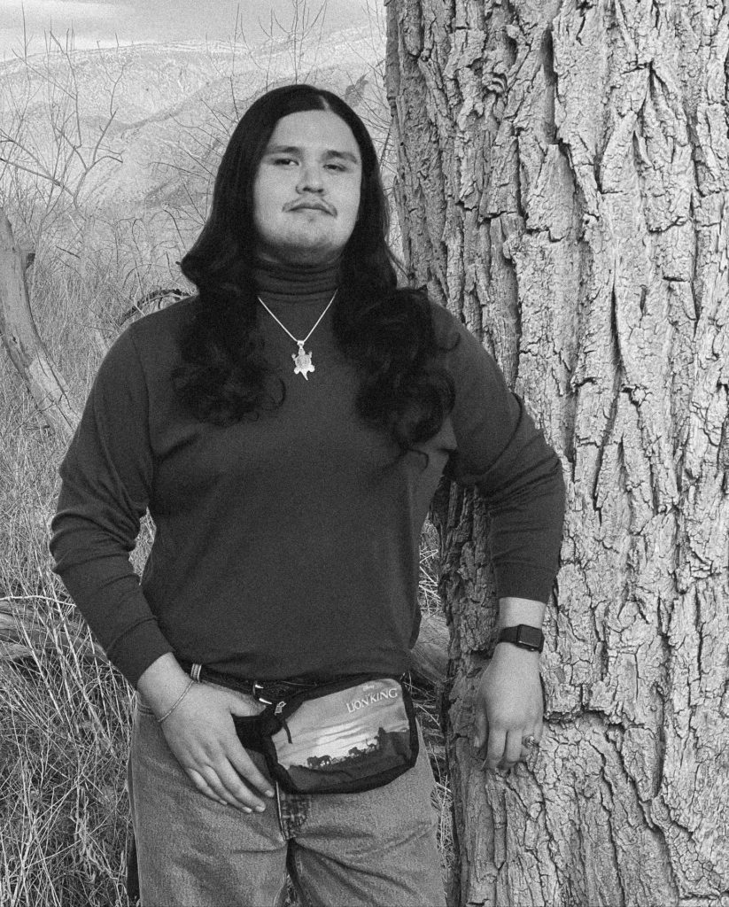 A black and white photo of an Indigenous man from the thighs up. He is wearing a black long sleeved shirt and leaning against a tree.