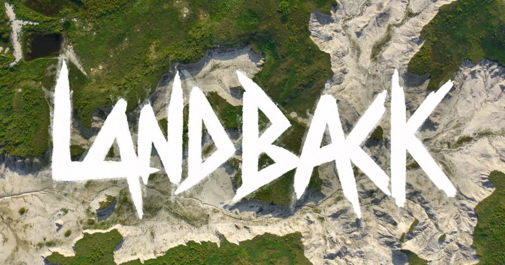 The word LANDBACK drawn over an aerial photo of land with green plateaus and mountains or creek beds.